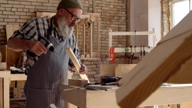 Medium shot of elderly carpenter with beard and trendy spectacles standing at workbench, drilling holes in plank using electrical drill
