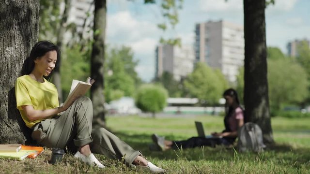 Asian female student sitting under tree leaning on trunk and reading book in park. Another college girl typing on laptop in background