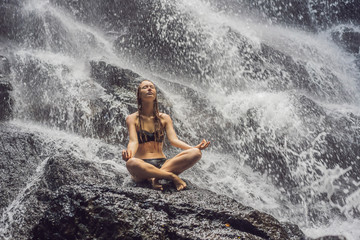 Wellness spa, vacation and yoga meditation concept. Young woman sitting in lotus position on the rock in tropical waterfall