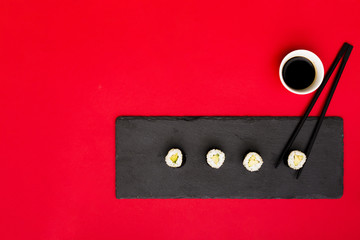 Row of asian rolls on slate plate with chopsticks and soya sauce over red backdrop