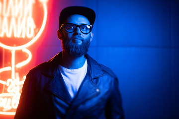 Hipster handsome man on the city streets being illuminated by neon signs. He is wearing leather biker jacket or asymmetric zip jacket with black cap, jeans and sunglasses.