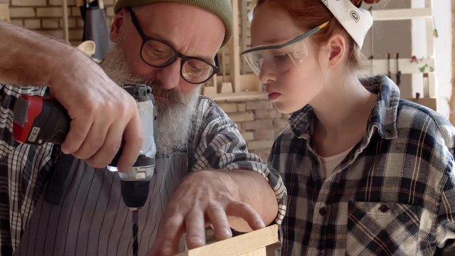 Senior trendy carpenter with beard and eyeglasses standing at workbench with his red-haired granddaughter and making holes in roof of wooden nesting box using drill