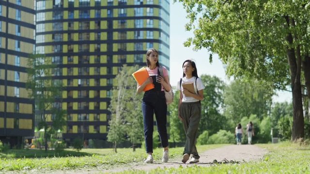Full length slow motion shot of two diverse female college students, Asian and Caucasian, with backpacks, textbooks and binders talking while walking home street after classes