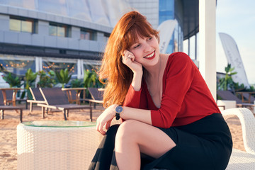 An attractive young girl with long curly redhead hair has bright red lipstick and freckles, wearing jumper and skirt, sitting on white chaise longue on terrace cafe by river. Sunny evening in summer