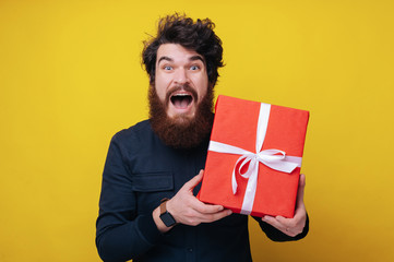 Handsome bearded man, looking excited at camera, holding a gift box, and standing over yellow...