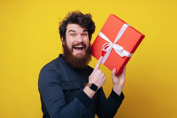 Handsome bearded man, looking excited at camera, holding a gift box, and standing over yellow...