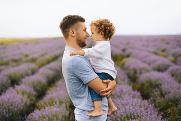 Photo of young father holding and hugging his little curly boy, having fun together, and posing in a lavender field