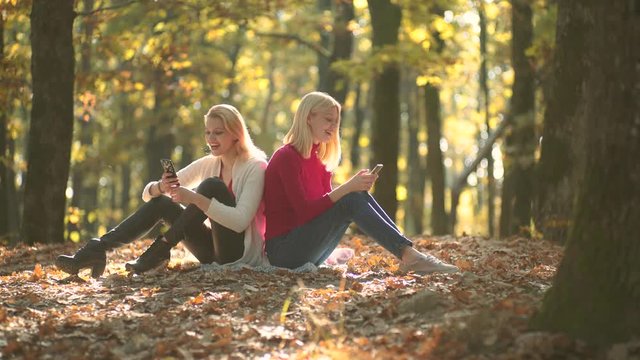 Two girls using smart phone. Warm Autumn sunny weather. Outdoor portrait of girls in autumn. Waiting in autumn park. Autumn girl.