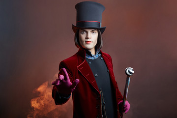 Fabulous circus man in a hat and a red suit posing in the smoke on a colored dark background. A...