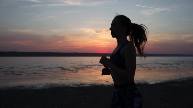 camera zooms out running woman silhouette in special suit enjoying cardio workout by calm sea at sunset slow motion
