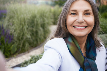Summertime, people, age and leisure concept. Close up of beautiful blue eyed mature European lady in elegant clothes enjoying nice summer day outdoors, taking selfie using cell phone, smiling happily