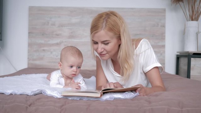 loving mother with book tells fairy tale story to baby daughter lying on the bed at home