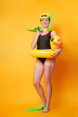 Photo of woman in black bathing suit in flippers with rubber ring in shape of duckling on orange background