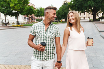 Image of charming young couple holding hands together while walking through city street with paper...