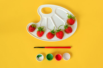 Red ripe strawberries on an artistic palette, brush and gouache on yellow background with copy...