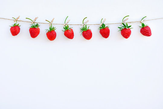Garland of red ripe strawberries hanging on wooden clothespin on rope twine, located on top edge on white wood background. Copy space Creative template for text, design, postcard or invitation