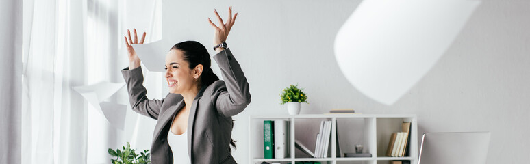 panoramic shot of irritated pregnant woman throwing papers in air while standing near table in office