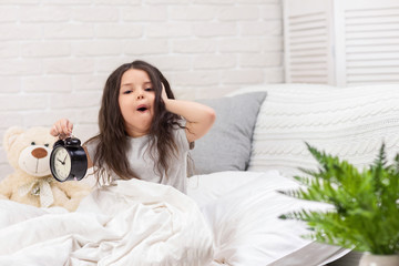 cute surprised little child girl in pyjamas with clock in bed. good morning