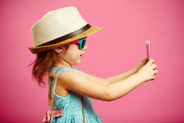 Beautiful little girl dressed in summer dress, wearing a straw hat and sunglasses, makes a selfie...
