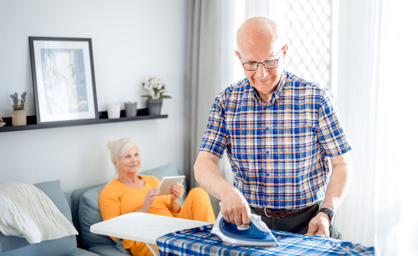 Senior man doing housework with iron and clothes while his wife using tablet - Powered by Adobe