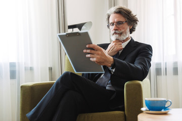 Portrait closeup of caucasian mature businessman holding clipboard while sitting on armchair in apartment