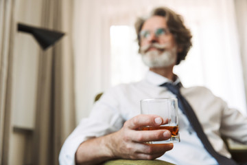 Handsome senior gray-haired business man sitting in chair indoors at home drinking alcohol.