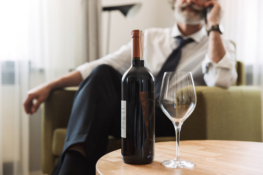 Image of attractive mature man talking on cellphone while sitting in bright apartment with bottle of wine - Powered by Adobe