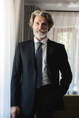 Bearded mature handsome grey-haired businessman posing indoors at home near window dressed in formal clothes.
