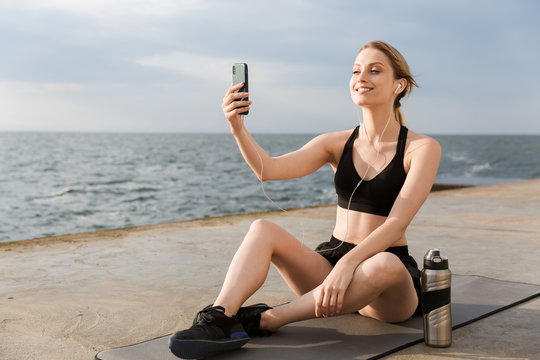 Image of positive woman using earphones and cellphone while sitting on mat near seaside in morning