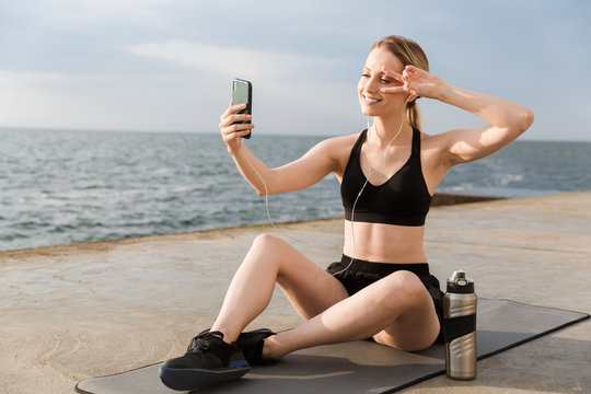 Image of positive woman taking selfie on cellphone and gesturing peace sign while sitting on mat near seaside