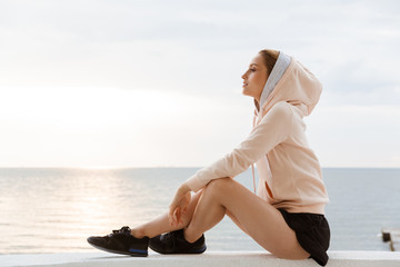 Image of calm woman looking aside while sitting on pier near seaside in morning