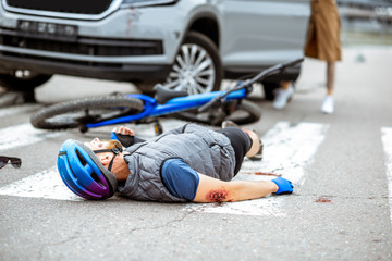 Obraz premium Road accident with injured cyclist lying on the pedestrian crossing near the broken bicycle and worried woman driver and car on the background