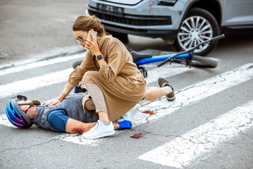 Road accident with injured cyclist lying on the pedestrian crossing near the broken bicycle, worried woman driver calling and checking men's pulse