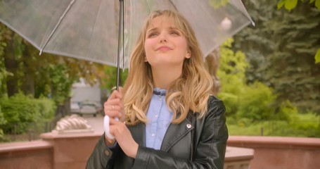 Pretty caucasian blonde student opens the umbrella preparing for rain being joyful in the green city park.