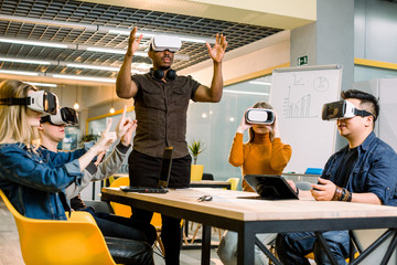Testing games. Young African man wearing virtual reality headset and gesturing while standing at the desk in creative office together with four multiethnical friends