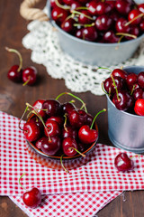 Composition with ripe cherries in metal bowls and clay brown plate. Vintage napkins as decor. Dacha style, countryside, cozy and cute, grandmother's kitchen. Dry flowers in vase on background