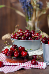 Composition with ripe cherries in metal bowls and clay brown plate. Vintage napkins as decor. Dacha style, countryside, cozy and cute, grandmother's kitchen. Dry flowers in vase on background