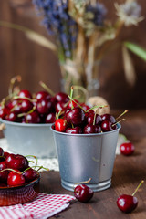 Composition with ripe cherries in metal bowls and clay brown plate. Vintage napkins as decor. Dacha style, countryside, cozy and cute, grandmother's kitchen. Dry flowers in vase on background