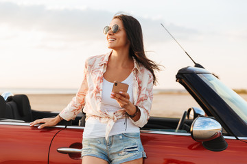 Happy smiling young pretty woman standing near car at the beach.