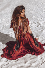 beautiful young fashionable woman in elegant dress sitting on sand at sunset