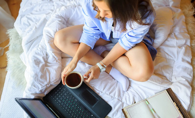 girl sits on the bed with a cup of morning coffee and works on a laptop, the work of a freelancer
