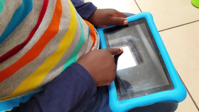 Black kid playing with a tablet and trying to switch it on. Toddler hands on tablet in kitchen .