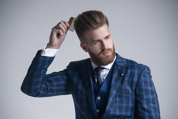 Confident in his perfect style. Confident young bearded man combing his hair and looking at away while standing against grey background. Dressed in full suit.