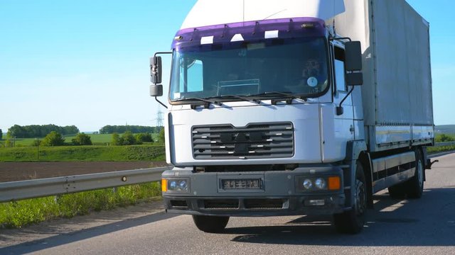 Camera follows to truck with cargo trailer driving on highway and transporting goods at summer day. Lorry riding through countryside with nature landscape at background. Slow mo Front view Close up