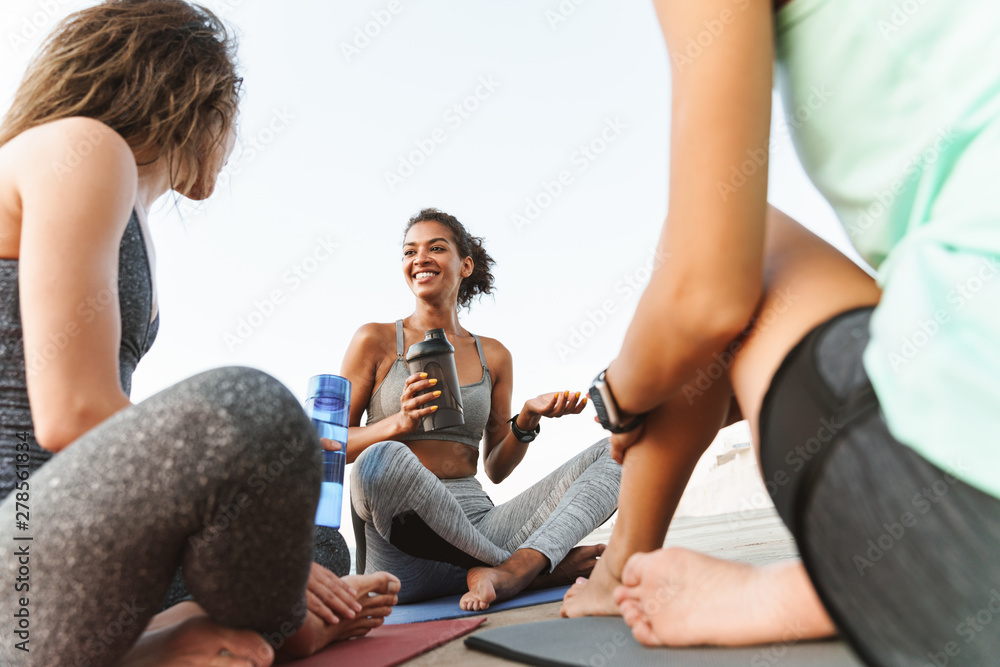 Sticker Photo of cheerful multiethnic sportswomen smiling and talking while sitting on yoga mats by seaside in morning