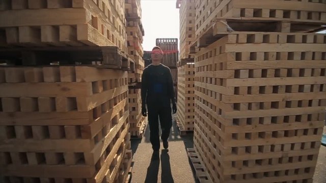 A young worker in uniform walks between wooden pallets in distribution outdoor warehouse. Slow motion
