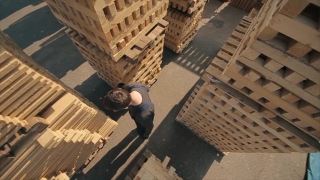 A young worker in uniform walks between wooden pallets in distribution outdoor warehouse. Slow motion. view from above