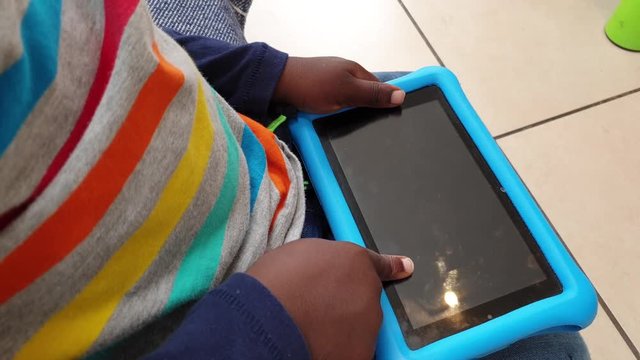 Black kid playing with a tablet and trying to switch it on. Toddler hands on tablet in kitchen.