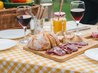 Party in the countryside. Table detail of a picnic decorated outdoors in summer with red wine in glasses, Italian sausage, homemade bread on a wooden cutting board on checkered tablecloth