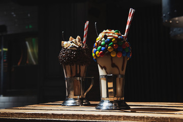 Homemade milkshakes with whipped cream colorful candies and chocolate filling on a wooden table. Black background. Sweetest desserts. Served in glass.